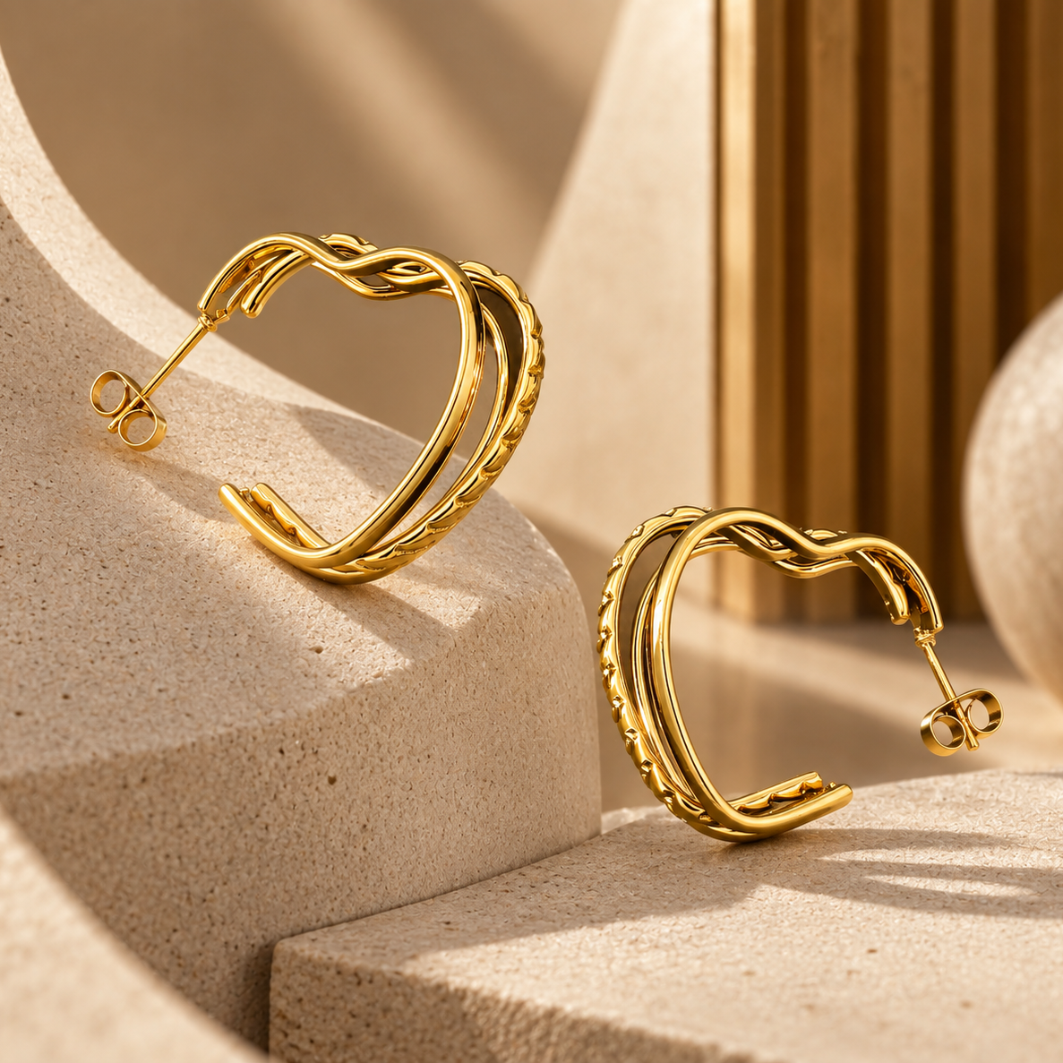 A pair of gold-toned twisted hoop earrings displayed on beige sculptural blocks in a warm, sunlit studio setting.